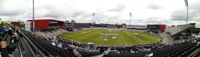 A large cricket stadium with partially filled stands surrounding a green field where players are practicing. Bright red and gray buildings are part of the stadium structure, and there are cloudy skies overhead.