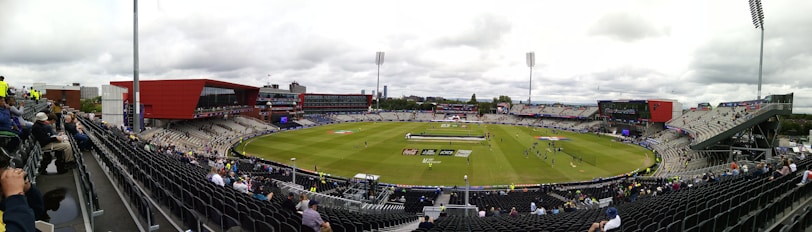 A large cricket stadium with partially filled stands surrounding a green field where players are practicing. Bright red and gray buildings are part of the stadium structure, and there are cloudy skies overhead.