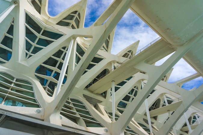 The image features the intricate and modern architectural design of a building with sweeping curves and pointed arches. The structure showcases a complex pattern of supports and windows with a light, airy appearance. The sky is a clear blue with scattered clouds, enhancing the overall striking look of the architecture.