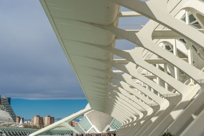 A series of modern, white architectural structures featuring sweeping lines and arches. The design creates a geometric and futuristic look. In the background, several tall buildings rise under a partially cloudy sky. Small groups of people can be seen walking in the distance, adding a sense of scale.