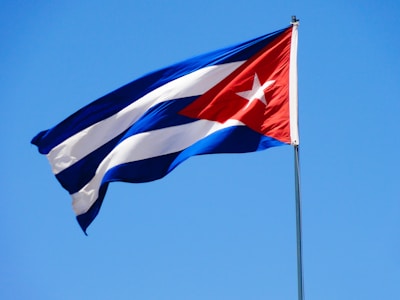 A Cuban flag waves in the wind against a clear blue sky, featuring a red triangle with a white star, and alternating blue and white stripes.