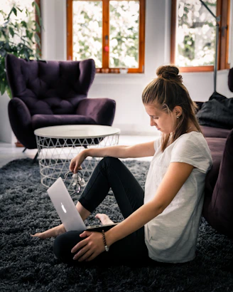 woman in white t-shirt and black leggings sitting on area rug while facing silver MacBook