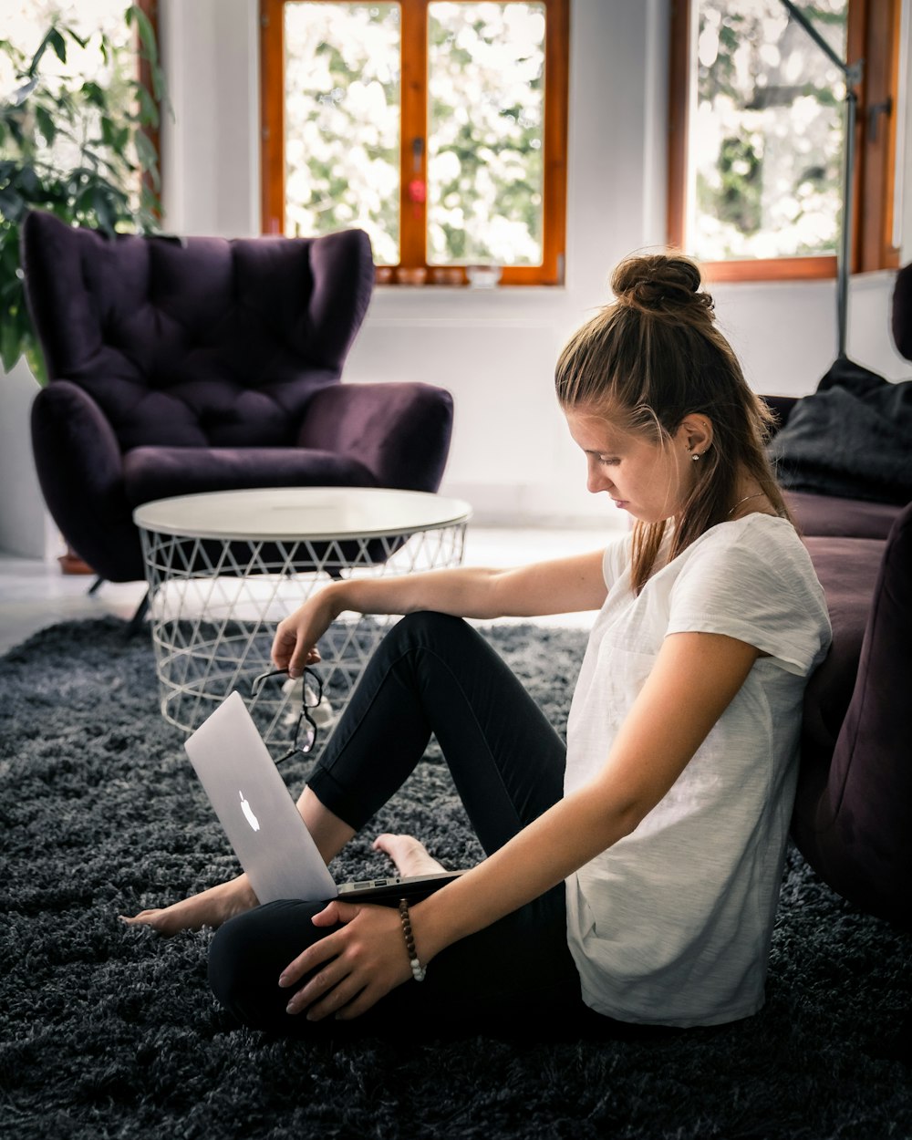 woman in white t-shirt and black leggings sitting on area rug while facing silver MacBook