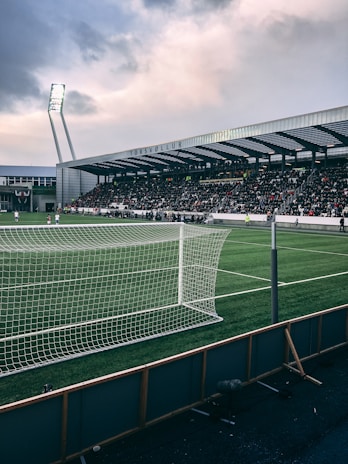 A football stadium filled with spectators under a cloudy sky. The field is green, with a white goalpost visible in the foreground. A well-illuminated stand is seen across the pitch.