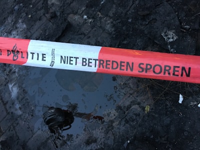 A red and white police tape is stretched across a wet, dark surface with a reflective puddle. The text on the tape reads 'POLITIE NIET BETREDEN SPOREN', indicating a restricted area.