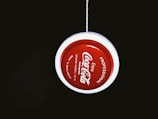 A red and white yo-yo with 'Coca-Cola' branding is suspended by a string against a dark background. The text around the yo-yo includes 'Professional' and 'Made in Bangladesh'.