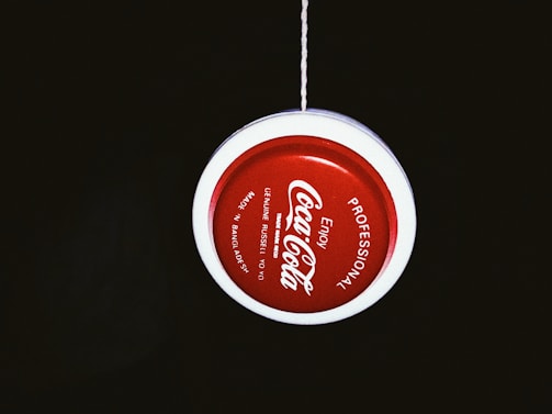A red and white yo-yo with 'Coca-Cola' branding is suspended by a string against a dark background. The text around the yo-yo includes 'Professional' and 'Made in Bangladesh'.