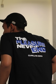 A person wearing a black t-shirt with a slogan on the back that reads 'The Pleasure Never Ends' in large, bold letters with a mix of white and blue colors. The shirt also displays 'CLAVE.LTD ©2019' in smaller text. The individual is also wearing a black cap.