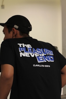 A person wearing a black t-shirt with a slogan on the back that reads 'The Pleasure Never Ends' in large, bold letters with a mix of white and blue colors. The shirt also displays 'CLAVE.LTD ©2019' in smaller text. The individual is also wearing a black cap.