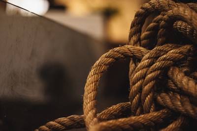 A close-up view of a thick, coiled rope with a textured surface, set against a blurred background. The lighting casts a warm tone over the rope, highlighting its fibers and intricate twists.