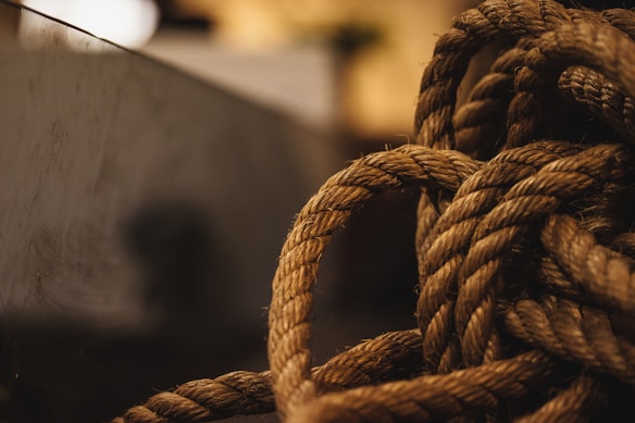 A close-up view of a thick, coiled rope with a textured surface, set against a blurred background. The lighting casts a warm tone over the rope, highlighting its fibers and intricate twists.