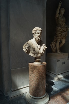 A marble bust of a bearded man with intricate carving stands on a polished cylindrical granite pedestal. Sunlight gently illuminates the bust, casting soft shadows. In the background, a larger statue of a standing figure is partially visible, also made of marble, with detailed drapery.