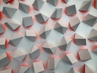 A repetitive pattern of three-dimensional geometric shapes projected outward against a background with soft gradients. The shapes appear to be cubes and pyramids, casting subtle shadows and reflections. The color palette consists of muted grays with hints of reddish tones between the shapes.