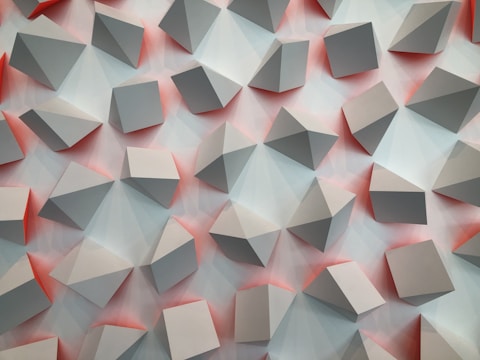 A repetitive pattern of three-dimensional geometric shapes projected outward against a background with soft gradients. The shapes appear to be cubes and pyramids, casting subtle shadows and reflections. The color palette consists of muted grays with hints of reddish tones between the shapes.