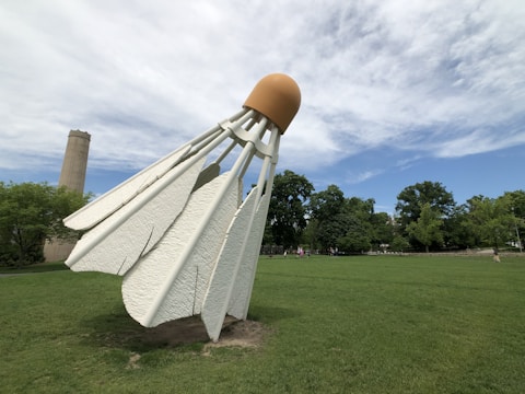 A large outdoor sculpture of a badminton shuttlecock placed on a grassy area. The shuttlecock is oversized and prominently displayed against a backdrop of trees and a partly cloudy sky. A tall, cylindrical structure is visible in the distance to the left.