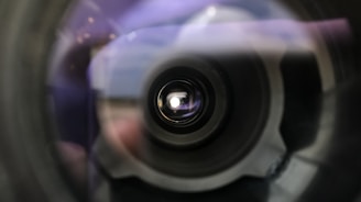 A close-up view of a camera lens with a focus on the center, showing the intricate details of the lens structure. The image contains blurred edges, adding a sense of depth and focus towards the center.