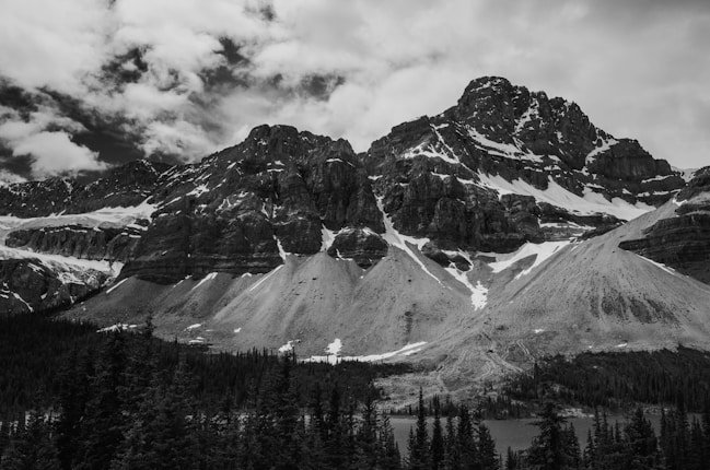 A rugged mountain landscape with snow-capped peaks and dramatic rock formations. Pine trees are clustered at the base, and the sky is filled with clouds.