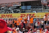 A group of people, many wearing orange apparel, are gathered at a fence, looking towards a large building with reflective windows displaying a DHL logo. The crowd appears excited and engaged, some are taking photos or videos.