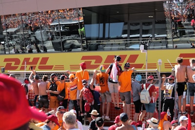 A group of people, many wearing orange apparel, are gathered at a fence, looking towards a large building with reflective windows displaying a DHL logo. The crowd appears excited and engaged, some are taking photos or videos.