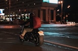 A person riding a motorcycle on a city street at night, carrying a large red backpack. The street is brightly lit by streetlights and the glow from nearby buildings, with some cars visible in the distance.