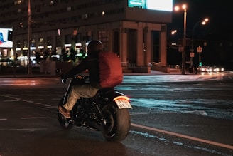 A person riding a motorcycle on a city street at night, carrying a large red backpack. The street is brightly lit by streetlights and the glow from nearby buildings, with some cars visible in the distance.