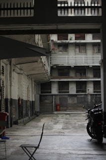 An empty courtyard surrounded by tall, weathered buildings with peeling paint. The area appears deserted, with a single folding chair and a parked motorcycle visible. The buildings feature barred windows and balconies, with a worn and industrial aesthetic.