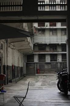 An empty courtyard surrounded by tall, weathered buildings with peeling paint. The area appears deserted, with a single folding chair and a parked motorcycle visible. The buildings feature barred windows and balconies, with a worn and industrial aesthetic.