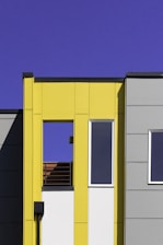 A modern building facade with a minimalistic design features bold geometric forms. The structure is characterized by flat surfaces and sharp lines, with a bright yellow panel contrasting against gray sections. Large rectangular windows provide a sleek and contemporary appearance, while the clear blue sky enhances the vibrant color palette.