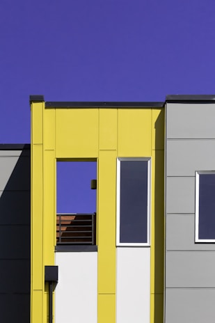 A modern building facade with a minimalistic design features bold geometric forms. The structure is characterized by flat surfaces and sharp lines, with a bright yellow panel contrasting against gray sections. Large rectangular windows provide a sleek and contemporary appearance, while the clear blue sky enhances the vibrant color palette.