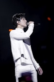 A person is performing on stage, passionately singing into a microphone. Dressed in a stylish white outfit, they are in profile view, with a focused expression. The background is dark, emphasizing the spotlight on the performer.