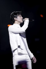 A person is performing on stage, passionately singing into a microphone. Dressed in a stylish white outfit, they are in profile view, with a focused expression. The background is dark, emphasizing the spotlight on the performer.