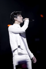 A person is performing on stage, passionately singing into a microphone. Dressed in a stylish white outfit, they are in profile view, with a focused expression. The background is dark, emphasizing the spotlight on the performer.