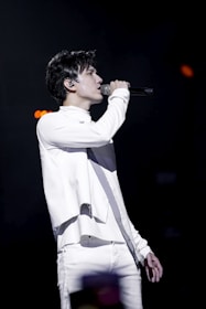 A person is performing on stage, passionately singing into a microphone. Dressed in a stylish white outfit, they are in profile view, with a focused expression. The background is dark, emphasizing the spotlight on the performer.