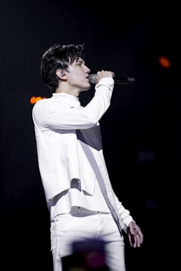 A person is performing on stage, passionately singing into a microphone. Dressed in a stylish white outfit, they are in profile view, with a focused expression. The background is dark, emphasizing the spotlight on the performer.