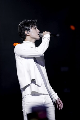 A person is performing on stage, passionately singing into a microphone. Dressed in a stylish white outfit, they are in profile view, with a focused expression. The background is dark, emphasizing the spotlight on the performer.