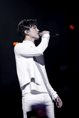 A person is performing on stage, passionately singing into a microphone. Dressed in a stylish white outfit, they are in profile view, with a focused expression. The background is dark, emphasizing the spotlight on the performer.