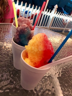 Colorful shaved ice treats in white cups, each with a different vibrant hue and a straw sticking out. The background displays a white picket fence and an outdoor setting, creating a casual snack atmosphere.