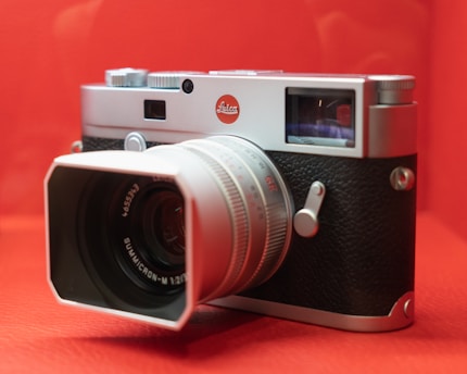 A silver and black camera with a large lens and a prominent logo is placed against a bright red background. The camera has a classic, vintage design with manual controls and a textured grip on the body.