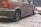 A person is using an air pump to inflate the rear left tire of a car. The vehicle appears to be worn and has visible dirt and scratches on its body. The scene is set outdoors on a dirt surface.