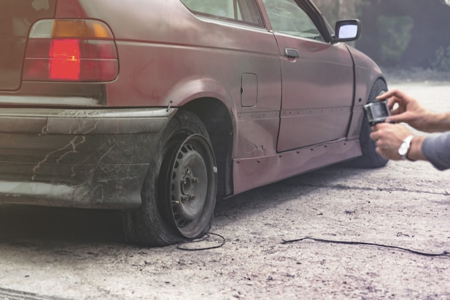 A person is using an air pump to inflate the rear left tire of a car. The vehicle appears to be worn and has visible dirt and scratches on its body. The scene is set outdoors on a dirt surface.