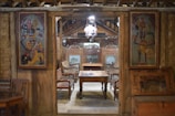 A traditional wooden interior with ornate wall panels and carved furniture. The room has wooden chairs and a central wooden table. Two decorative paintings with vibrant colors are mounted on either side of the entrance.