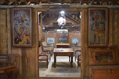 A traditional wooden interior with ornate wall panels and carved furniture. The room has wooden chairs and a central wooden table. Two decorative paintings with vibrant colors are mounted on either side of the entrance.