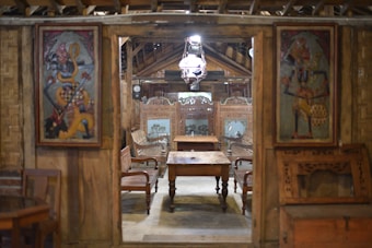 A traditional wooden interior with ornate wall panels and carved furniture. The room has wooden chairs and a central wooden table. Two decorative paintings with vibrant colors are mounted on either side of the entrance.