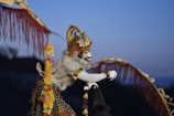 A person dressed in elaborate traditional Balinese attire with a striking white face paint, adorned with a gold crown and decorative elements. The costume includes intricate patterns and a majestic cape, set against a twilight sky.
