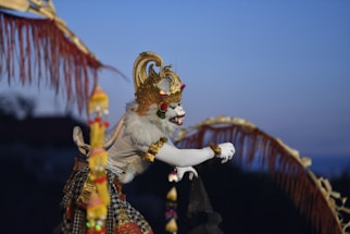 A person dressed in elaborate traditional Balinese attire with a striking white face paint, adorned with a gold crown and decorative elements. The costume includes intricate patterns and a majestic cape, set against a twilight sky.