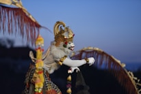 A person dressed in elaborate traditional Balinese attire with a striking white face paint, adorned with a gold crown and decorative elements. The costume includes intricate patterns and a majestic cape, set against a twilight sky.