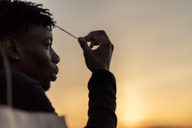 A person is holding a strand of their hair in silhouette against a golden sunset. The background features a soft gradient of warm colors transitioning from orange to yellow, creating a serene and contemplative atmosphere.