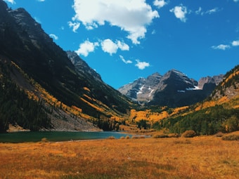 A picturesque landscape featuring majestic, snow-capped mountains beneath a bright blue sky with scattered clouds. Vibrant autumn foliage in shades of yellow and orange covers the slopes, contrasting with lush green pine forests. A tranquil lake sits at the base of the mountains, surrounded by a field of golden grass.