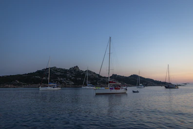 A classic sailboat anchored near a rocky coastline at sunset.
