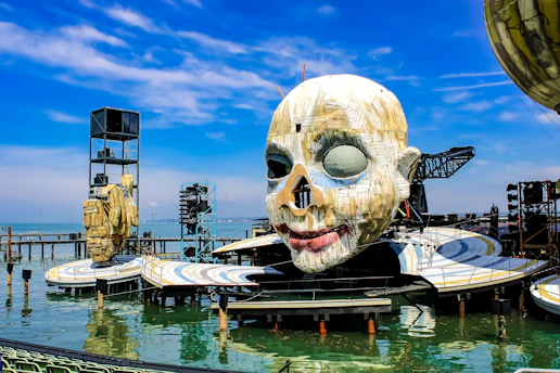 A dramatic outdoor stage setup on water, featuring a massive, artistic sculpture of a face with a painted smile and one eye covered. The stage appears to have walkways and platforms, with additional mechanical structures and set pieces around. The backdrop is a clear blue sky and calm waters.